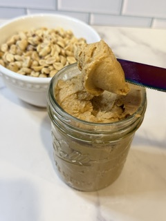 Homemade peanut butter in a glass far with a bowl of roasted peanuts in the background
