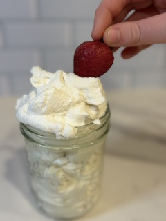 Homemade whipped cream in a glass jar being scooped with a fresh strawberry, showing creamy homemade whipped cream ready to serve.